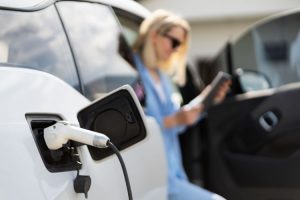 A car with an electric charger cable plugged into its charger socket, and with a woman in the background